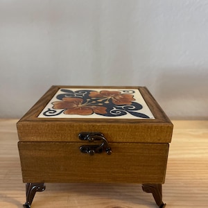 May include: A rectangular wooden box with a hinged lid featuring a ceramic tile inlay. The tile displays a painted floral pattern in shades of orange and dark blue. The box has a metal latch closure and stands on four decorative metal feet.