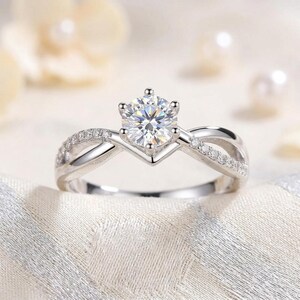 May include: A silver-toned ring featuring a central round-cut clear gemstone held by a six-prong setting. The band has a crossover design with small pavé-set clear stones embedded along the upper shoulders of the metal shank.