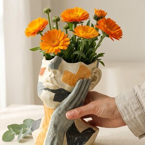 Peut inclure: Un vase en céramique en forme de tête avec un motif peint abstrait et texturé dans des tons orange, gris et noir. Le vase représente un visage avec une main posée sur la joue et contient un bouquet de fleurs de souci orange vif.