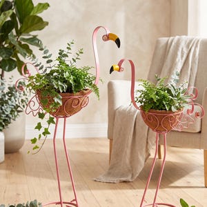 Op de afbeelding: Twee roze metalen plantenstandaards in de vorm van een flamingo met lange poten en gekrulde staartdetails. Elke flamingo heeft een geel met zwarte snavel en bevat een ronde bruine kokosvezel voering gevuld met groene planten.
