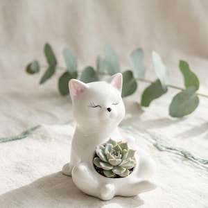 Peut inclure: Un cache-pot en céramique blanche en forme de chat avec une finition brillante, des oreilles teintées de rose et des yeux fermés. Le chat est assis et une petite plante succulente vert clair est placée dans une ouverture circulaire sur son corps.