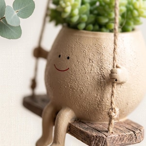 Peut inclure: Un pot de fleurs en céramique beige à la texture granuleuse, en forme de personnage rond au visage souriant, assis sur une petite balançoire en bois suspendue par des cordes en jute. Il contient une plante succulente verte.