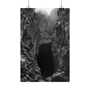 Peut inclure: Une impression d'art mural en noir et blanc représentant l'entrée d'une grotte profonde et sombre nichée entre des falaises rocheuses escarpées. La composition verticale met en valeur les textures de pierre et un ciel brumeux au sommet.