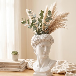 Op de afbeelding: Een witte keramische vaas in de vorm van een klassieke mannelijke buste, met gedetailleerde gelaatstrekken en krullend haar. De vaas is gevuld met groene eucalyptus takken en beige pampasgras pluimen voor een textuurrijk geheel.