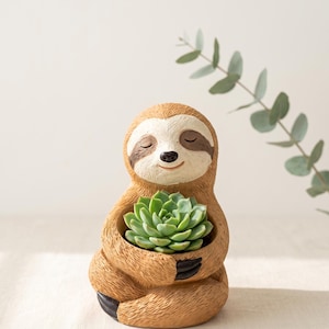 May include: A brown sloth-shaped planter with a textured fur finish, featuring a cream-colored face with closed eyes and a small black nose. The sloth is sitting in a cross-legged position, holding a green succulent plant in its arms.