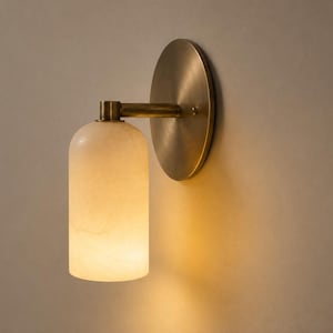 May include: A wall sconce featuring a cylindrical, translucent white stone shade that emits a warm glow. The fixture includes a circular, brushed brass backplate and a horizontal arm that connects the shade to the wall-mounted base.