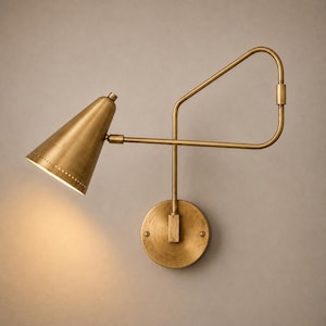 Puede incluir: Una lámpara de pared con acabado en latón cepillado, que presenta una pantalla cónica con un borde perforado y un brazo ajustable. La lámpara tiene una placa base circular y un marco geométrico que se extiende para sostener la luminaria.