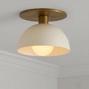 Könnte beinhalten: Eine Deckenleuchte mit einem matten, cremeweißen, kuppelförmigen Schirm und einem runden, messingfarbenen Metalldeckenanschluss. Die Leuchte enthält ein zentrales, leuchtendes Leuchtmittel, das innerhalb der Kuppel sichtbar ist und sanftes Licht abgibt.