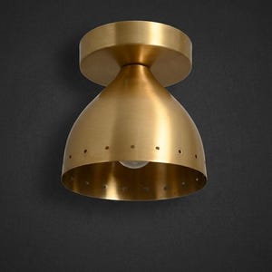 Könnte beinhalten: Eine Deckenleuchte aus Messing mit einem glockenförmigen Schirm. Der Schirm hat eine glatte, gebürstete Metalloberfläche und eine einzelne Reihe kleiner, gleichmäßig verteilter kreisförmiger Perforationen entlang der unteren Kante.