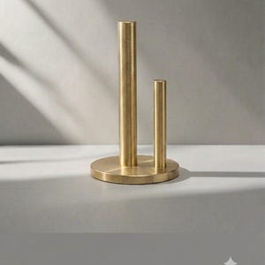 May include: A brushed brass metal stand featuring a circular base with two vertical cylindrical rods of unequal heights attached to the top surface. The taller rod is positioned on the left, while the shorter rod is located on the right side.