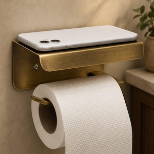 May include: A brushed brass toilet roll holder with an integrated flat shelf on top. A white smartphone rests horizontally on the shelf, while a standard roll of white toilet paper hangs from the horizontal bar underneath the metal fixture.