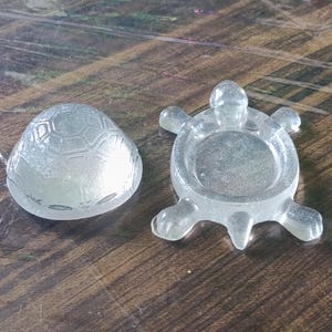 Puede incluir: Un joyero de resina transparente y brillante con forma de tortuga, compuesto por dos piezas separadas. La tapa presenta un patrón de caparazón hexagonal moldeado, mientras que la base tiene forma de tortuga con cuatro aletas y cabeza.