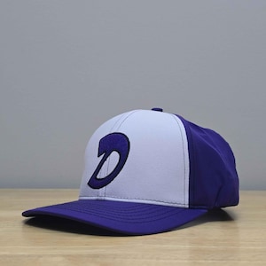 Puede incluir: Una gorra de béisbol con un panel frontal de color morado claro y una letra D bordada en morado oscuro. La visera, los paneles laterales y los paneles traseros son de un color morado oscuro sólido. La gorra tiene una corona estructurada.