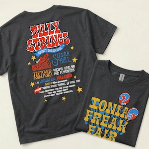 Può includere: Una t-shirt grigio scuro con una grafica di un concerto sul retro con il testo Billy Strings August 28 & 29, 2023, e una seconda t-shirt che mostra il testo Ionia Freak Fair in giallo e blu con due grafiche di palloncini rossi e blu.