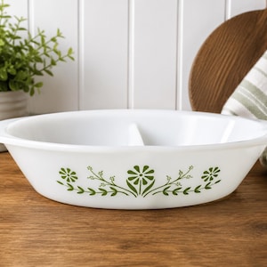 Peut inclure: Un plat de cuisson ovale divisé en verre blanc, orné d'un motif floral vert sur l'extérieur. Le plat comporte une séparation centrale qui divise l'intérieur en deux sections, avec des bords lisses et arrondis et une finition brillante.