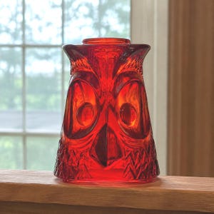 May include: A vibrant red glass owl-shaped votive holder featuring a moulded design with prominent circular eyes, a pointed beak, and textured feather patterns across the body. The translucent glass allows light to pass through the form.