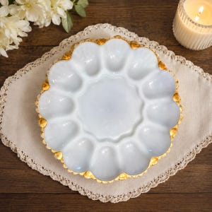 Op de afbeelding: Een ronde witte porseleinen eierschaal met twaalf individuele ovale inkepingen in een cirkelvormig patroon. De geschulpte buitenrand van de schaal is rondom afgewerkt met een metallic gouden rand.