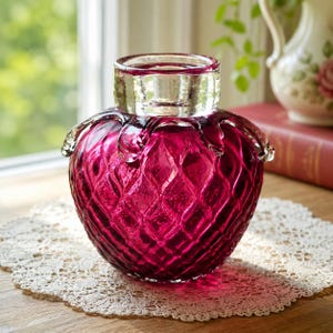 May include: A deep magenta glass vase with a rounded, bulbous body featuring a textured diamond pattern. The vase has a clear, smooth glass neck and a wide, circular opening at the top, with decorative glass drips extending from the rim.