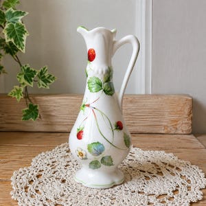 Könnte beinhalten: Eine weiße Keramik-Krugvase mit einem aufgemalten Muster aus roten Erdbeeren, grünen Blättern und kleinen weißen Blüten. Das Gefäß hat einen schlanken Hals, einen geschwungenen Henkel, einen gewellten Rand und einen ausgestellten Fuß mit grünem Rand.