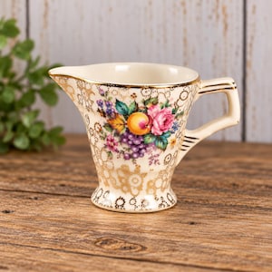 Peut inclure: Un pot à crème en céramique de couleur crème orné d'un motif central de fleurs et de fruits comprenant des roses roses, des raisins violets et une pêche orange. L'extérieur présente un motif en filigrane doré et un bord et une anse dorés.