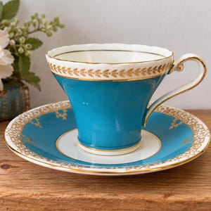 May include: A teal porcelain teacup and saucer set featuring a white interior and a gold leaf pattern along the rim and base. The teacup has a curved handle and a scalloped edge, while the saucer displays a matching gold filigree design.