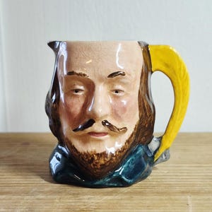 Peut inclure: Une cruche en céramique en forme de tête humaine avec un visage détaillé, des cheveux bruns et une moustache. L'objet présente une finition brillante avec un fin craquelé visible, un col bleu sarcelle et une anse jaune vif en forme de feuille.