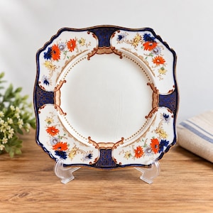 May include: A square ceramic plate with scalloped edges featuring a white center, deep blue patterned borders, and ornate gold-toned scrollwork. The rim is decorated with vibrant orange, yellow, and blue floral motifs on a white background.