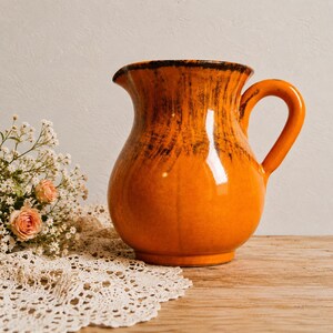 May include: A glossy orange ceramic pitcher with a rounded body and a curved handle. The upper portion of the pitcher features dark brown, vertical drip-like glaze accents that contrast with the vibrant orange finish of the lower section.