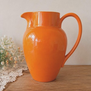 Puede incluir: Una jarra de cerámica de color naranja brillante con un acabado liso y lustroso. El recipiente presenta un cuerpo redondeado, un pico vertedor ancho y un asa curva que se extiende desde el lateral. El diseño es sencillo y funcional.