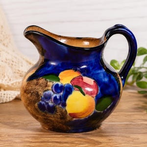 May include: A ceramic pitcher featuring a deep blue glaze with a hand-painted fruit motif. The design includes clusters of purple grapes, yellow peaches, and a red apple against a textured brown base. The pitcher has a curved handle and a wide spout.