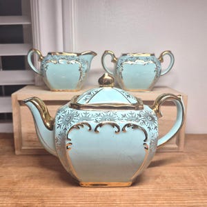 Pode incluir: Um conjunto de chá de porcelana azul-claro composto por um bule com tampa, uma leiteira e um açucareiro. Cada peça apresenta bordas douradas, padrões florais dourados e um acabamento de esmalte craquelado na superfície de cerâmica.