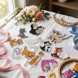 Könnte beinhalten: Eine Sammlung von gestickten Aufnähern mit verschiedenen animierten Katzenfiguren, darunter eine rosa-lila gestreifte Katze, eine schwarz-weiße Katze und mehrere orangefarbene Kätzchen, angeordnet auf einem weißen Stoff mit einem goldenen Diadem und Blumen.