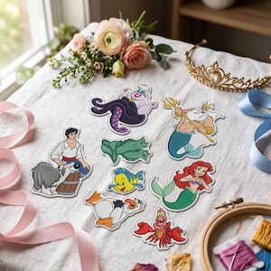 May include: A collection of embroidered patches featuring characters from The Little Mermaid, including Ariel, King Triton, Ursula, Flounder, Sebastian, Scuttle, Prince Eric, Max the dog, and two eels, arranged on a white fabric surface.