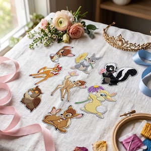 May include: A collection of embroidered fabric patches featuring various cartoon forest animals, including a fawn, a rabbit, a skunk, and an owl. The patches are arranged on a white fabric surface alongside a gold tiara and pink ribbons.