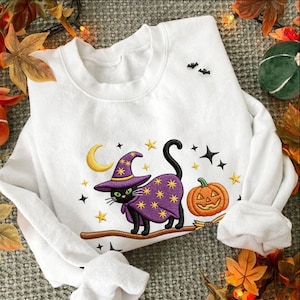 Op de afbeelding: Een wit sweatshirt met lange mouwen en een borduursel van een zwarte kat met een paarse heksenhoed en cape met gele sterren. De kat staat op een bruine tak naast een oranje pompoen, een gele halve maan en kleine zwarte vleermuizen.