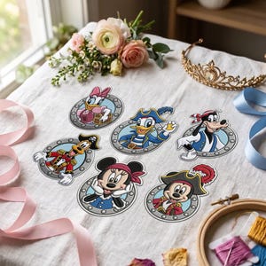 Puede incluir: Seis parches bordados con personajes de Disney en trajes de pirata, cada uno enmarcado por un diseño circular de ojo de buey gris. El conjunto incluye a Daisy, Donald, Goofy, Minnie y Mickey, dispuestos sobre una superficie de tela blanca.