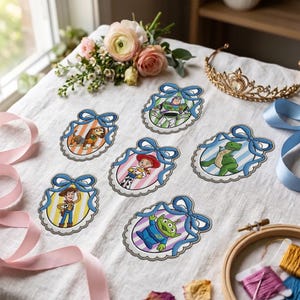 Könnte beinhalten: Sechs gestickte Aufnäher mit Toy Story-Figuren, darunter Woody, Buzz Lightyear, Jessie, Rex, Slinky Dog und ein Alien. Jede Figur ist in einem Oval mit einer blauen Schleife oben eingerahmt, platziert auf einem weißen Leinenstoffhintergrund.