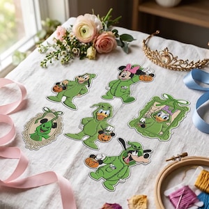 Może przedstawiać: Kolekcja sześciu wycinanych naklejek przedstawiających postacie Disneya ubrane w zielone kostiumy Oogie Boogie na Halloween. Naklejki zawierają Goofy'ego, Myszkę Minnie, Daisy Duck i Donalda Ducka, niektóre w ozdobnych ramkach.