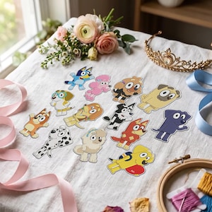 Op de afbeelding: Een verzameling cartoonhondstickers met verschillende rassen en kleuren, waaronder blauwe, gele, roze en gevlekte ontwerpen. De stickers zijn gerangschikt op een witte stoffen ondergrond naast een gouden tiara, roze lint en een borduurring.