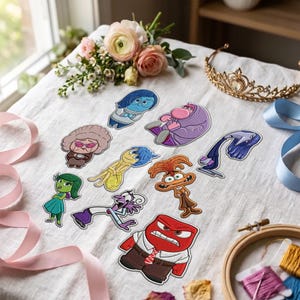 May include: A collection of embroidered patches featuring various colorful cartoon characters from the movie Inside Out, including Sadness, Joy, Anger, Disgust, Fear, and others, arranged on a white fabric surface with ribbons and a tiara.