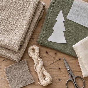 May include: A craft supply set featuring folded beige and olive green textured fabrics, a white felt Christmas tree cutout, a square of grey felt, a skein of cream embroidery thread with a needle, four straight pins, and metal scissors.