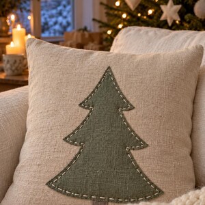 May include: A square scatter cushion made of light beige textured fabric featuring a centred dark olive green fabric cutout in the shape of a pine tree. The tree is attached to the cushion cover with visible white running stitch embroidery.