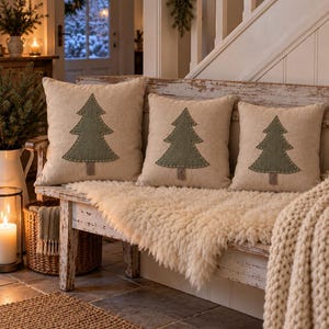 May include: Three square beige throw pillows arranged in a row, each featuring a centred olive green fabric Christmas tree appliqu&eacute; with a brown trunk and visible white decorative stitching along the edges of the tree shape.