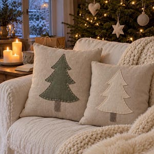 May include: Two square scatter cushions made of light beige fabric, each featuring a central Christmas tree applique. One cushion displays a dark green tree with white stitched edges, while the other features a cream-coloured tree with matching stitching.