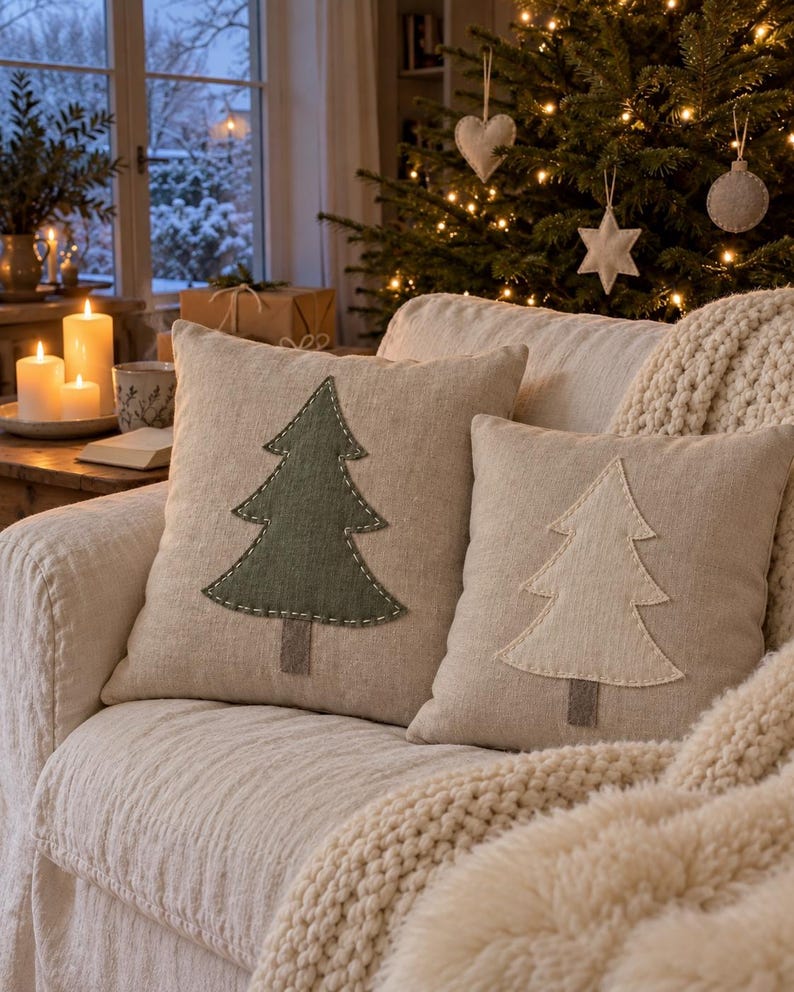 May include: Two square scatter cushions made of light beige fabric, each featuring a central Christmas tree applique. One cushion displays a dark green tree with white stitched edges, while the other features a cream-coloured tree with matching stitching.