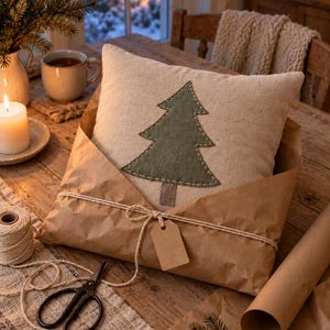 May include: A square beige throw pillow featuring a centred dark green fabric Christmas tree applique with visible white stitching, wrapped in brown kraft paper and tied with white twine, including a blank rectangular paper gift tag.