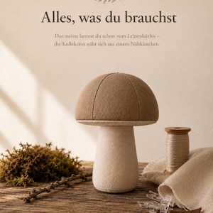 Puede incluir: Un alfiletero con forma de seta con un sombrero de tela marrón y un tallo de tela color crema. La imagen incluye el texto KAPITEL EINS, Alles, was du brauchst, y Das meiste kennst du schon vom Leinenkürbis – die Kollektion näht sich aus einem Nähkästchen.