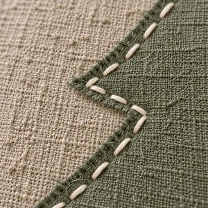 May include: A textured fabric surface featuring a beige section joined to an olive green section with a zigzag seam. The seam is reinforced with visible, thick white running stitches that follow the jagged edge of the green fabric panel.