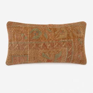 Puede incluir: Un cojín lumbar rectangular con una funda de tejido textil en tonos óxido, terracota y verde salvia apagado. La tela muestra un patrón geométrico intrincado y descolorido con una textura visible y un aspecto desgastado en toda la superficie.