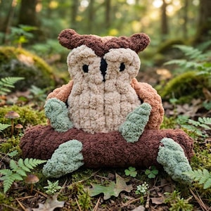 Puede incluir: Un juguete de peluche con forma de búho hecho de hilo suave y texturizado en tonos marrón, beige y verde salvia. El búho presenta un cuerpo redondeado, mechones en las orejas y ojos oscuros, posado sobre una base cilíndrica marrón.